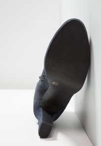 Semelle noire d'une botte à talon haut en daim bleu foncé, taille 37, appuyée contre un mur blanc sur un sol blanc.