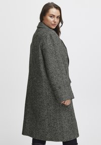 Cappotto a motivo a spina di pesce in bianco e nero con un design strutturato, colletto a punte e maniche lunghe, mostrato di lato.