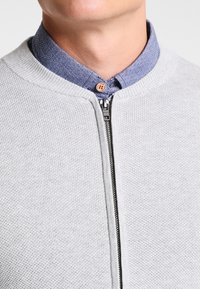 Lichtgrijze gebreide trui met ritssluiting, een geribde kraag en een spijkerbroekshirt eronder, met een zichtbare knoop en subtiele textuur.