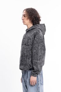 Jovem com cabelo cacheado, vestindo um moletom preto desbotado com letras em patch e jeans claros largos, de perfil contra um fundo branco.