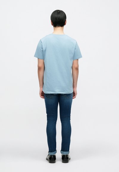Ανοιχτό γαλάζιο βαμβακερό μπλουζάκι με κοντά μανίκια, σε συνδυασμό με σκούρο τζιν και μαύρα παπούτσια, φαίνεται από πίσω.