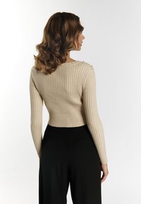 Beige geripptes Strickpullover mit langen Ärmeln und tailliertem Schnitt, mit Knopfdetails an der Schulter. Kombiniert mit schwarzen Hosen.