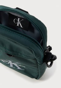 Grønn nylonpose med glidelåslukking, med svart logoetikett inni og svart metallaccent, glatt tekstur og kompakt design.