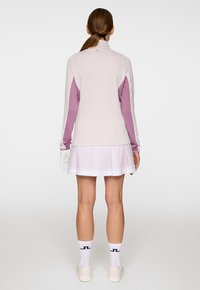 Kvinna som står vänd bort med en rosa och lavendelfärgad jacka, ljusrosa plisserad kjol, vita handskar, strumpor och sneakers.