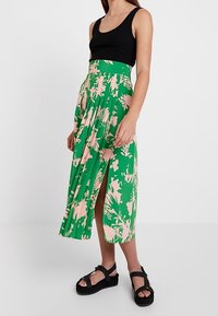 Grüner Blumenkleid mit pinken Akzenten, plissierter Schnitt, hohe Taille und seitlicher Schlitz. Kombiniert mit einem schwarzen Tanktop und klobigen schwarzen Sandalen.