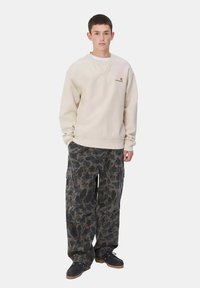Sudadera beige con un logotipo en el pecho, combinada con pantalones cargo de camuflaje oscuros con bolsillos. Zapatos negros con suela marrón.