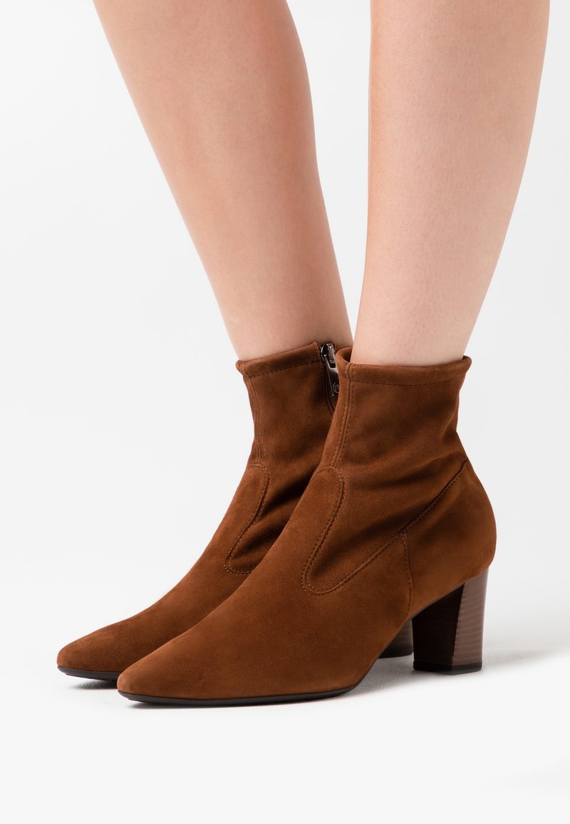 Bottines en daim marron à talons blocs portées sur jambes nues, avec fermeture éclair latérale et bouts pointus sur fond blanc.