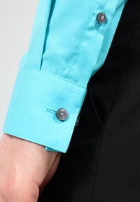 Lichtblauwe overhemdmanchet met grijze knopen, met een soepele katoenen textuur en hoekig ontwerp. Zwarte stof zichtbaar op de achtergrond.