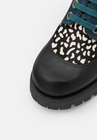 Chaussure en cuir noir avec semelle épaisse, lacets bleu sarcelle et panneau à imprimé léopard près de l'avant-pied sur fond blanc.