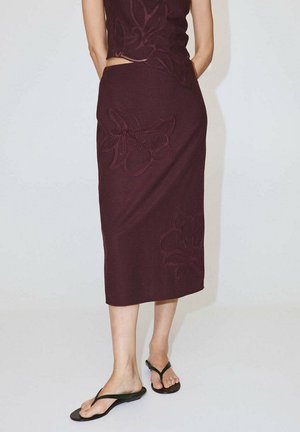 Personne portant une jupe midi violet foncé avec une broderie florale et des tongs noires, debout devant un fond clair uni.