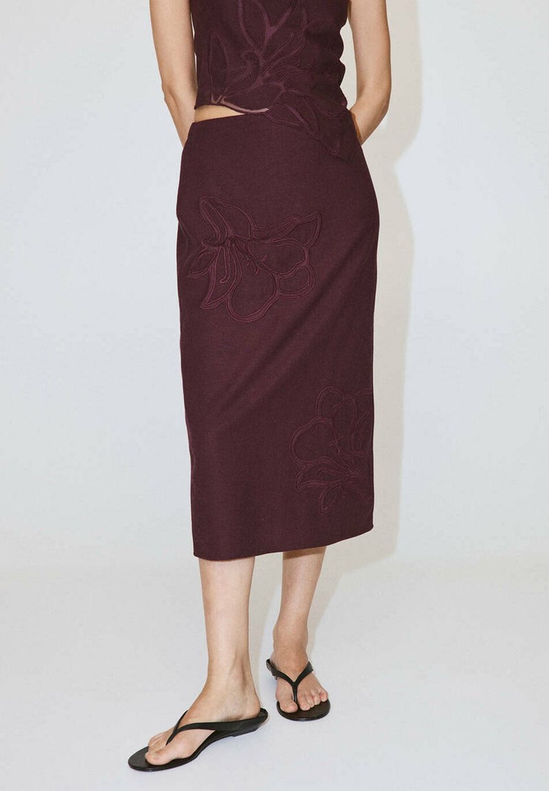 Personne portant une jupe midi violet foncé avec une broderie florale et des tongs noires, debout devant un fond clair uni.