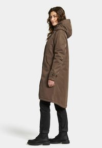 Bruine lange jas met een capuchon, voorzien van zijzakken en knoopmanchetten, gecombineerd met zwarte jeans en laarzen. Glamoureuze textuur.