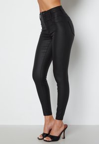 Žena oblečená v černých skinny kalhotách z umělé kůže vysokého pásu a černých otevřených lodičkách na vysokém podpatku, stojící na jednobarevném světlejším pozadí.