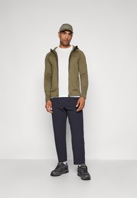 Oliven grøn zip-up jakke over en hvid t-shirt, kombineret med navy bukser og grå sneakers. Outfittet har et minimalistisk design og en baseball cap.