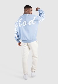 Lichtblauwe sweatshirt met witte cursieve tekst op de mouwen en de rug, geribbelde manchetten en zoom, gecombineerd met beige sweatpants en witte sneakers.