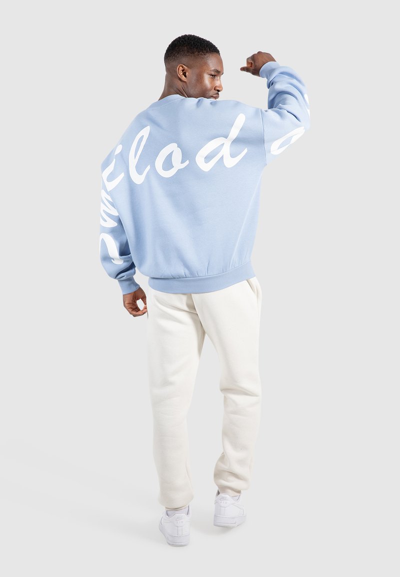 Lichtblauwe sweatshirt met witte cursieve tekst op de mouwen en de rug, geribbelde manchetten en zoom, gecombineerd met beige sweatpants en witte sneakers.