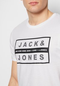 White cotton t-shirt featuring a black rectangular graphic text design with "JACK & JONES" and other text elements in a bold, contrasting font.
