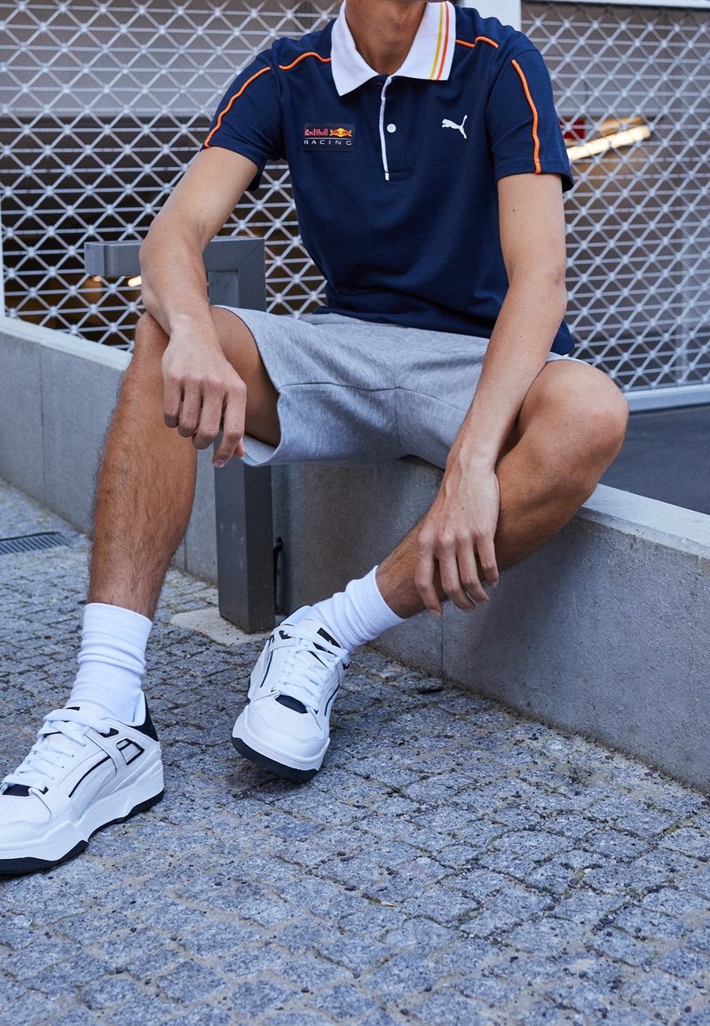 Polo shirt navy à rayures jaunes et oranges, shorts gris et baskets blanches avec accents noirs. Tenue décontractée présentée dans un cadre urbain.