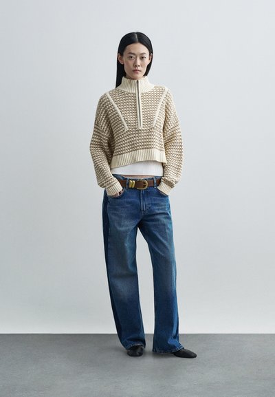 Pulover bej, tricotat, croială scurtă, cu guler înalt și model texturat. Asortat cu blugi albaștri, lejeri, și curea maro. Pantofi negri.