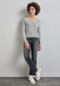 Grå V-ringad tröja som är parat med mörka jeans och vita sneakers. Tröjan har ribbade muddar och jeansen har subtila sömnadsdetaljer.