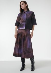 Camisa colorida de manga corta en tonos morados y negros, combinada con una larga falda plisada a juego. La modelo lleva botas de tacón negro altas.