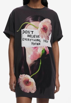 Czarny oversize'owy T-shirt z wizerunkiem kobiecej twarzy częściowo zakrytej rozerwanym papierem z napisem "NIE WIERZ WE WSZYSTKO, CO MYŚLISZ" oraz trzema różowymi kwiatami.