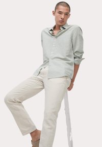 Camisa de lino verde claro con botones, mangas enrolladas y cuello, combinada con pantalones de lino beige y sandalias neutras, posando sobre un taburete.