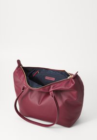 Sac fourre-tout bourgogne en matériau synthétique lisse avec fermeture à glissière, doté d'un intérieur bleu marine et de deux anses solides.
