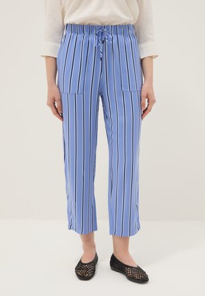 Donna che indossa pantaloni corti blu con righe verticali bianche e nere, tasche anteriori e scarpe basse nere intrecciate, in piedi su pavimento bianco.