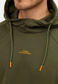 Sudadera color verde oliva hecha de una tela suave, con un bolsillo tipo canguro y el texto "ICONIC EXPERIENCE" en naranja, con detalles de cordón a juego.
