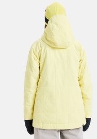 Gele pufferjack met een capuchon, voorzien van een gewatteerd design. Draagt een bijpassende gele beanie en zwarte handschoenen.