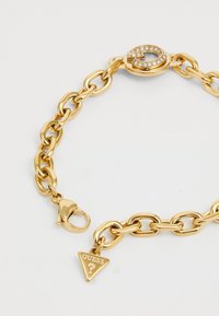 Pulsera de cadena de oro con eslabones grandes, adornada con un cierre decorativo y un charm triangular que lleva la palabra "GUESS."