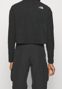 Personne portant une veste polaire noire raccourcie avec le logo The North Face sur le haut du dos et un pantalon noir assorti, vue de dos.