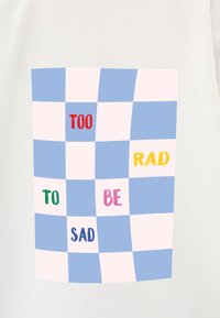 Koszulka t-shirt z jasnobeżowym tłem, na którym znajduje się niebieski i różowy wzór w kratę. Tekst zawiera słowa "TOO," "RAD," "TO," "BE" i "SAD" w różnych kolorach.