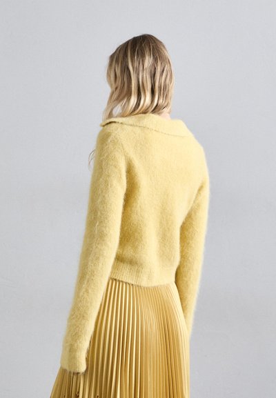 Κίτρινη χνουδωτή μπλούζα με στρογγυλή λαιμόκοψη, συνδυασμένη με μια κίτρινη φούστα με πτυχές. Η μπλούζα διαθέτει μακριά μανίκια και υφή.