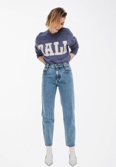 Lilla genser med stor hvit "DALL" tekst, kombinert med høytlivs lyse blå jeans. Modellen har på seg hvite spisse sko og ankelstrømper.