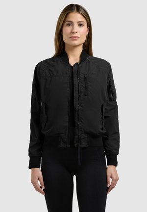 Woman standing wearing a black bomber jacket with zippered pockets, ribbed cuffs, and collar against a plain light background.