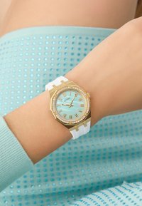 Gold watch with octagonal face, white rubber strap, light blue dial, and Roman numerals. Surrounded by a glittering crystal bezel.