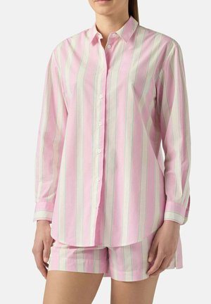 Donna che indossa una camicia a righe verticali rosa e bianche con maniche lunghe abbottonata e pantaloncini abbinati, in piedi davanti a uno sfondo neutro.