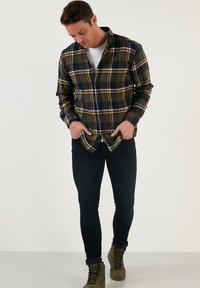 Cămașă din flanel de culoare verde închis, albastru marin și galben cu un model în carouri; cu nasturi în față, mâneci lungi, purtată peste un tricou alb și blugi slim-fit.