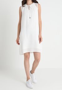 Biała sukienka bez rękawów z wiązaniem na szyi, koronkowymi akcentami na ramionach i luźnym krojem, zestawiona z białymi trampkami.