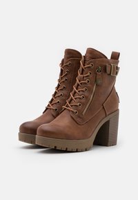 Paire de bottines en cuir marron à lacets, avec fermetures éclair latérales, sangles à boucle, et talons blocs épais sur fond blanc.