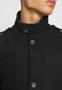 Zwarte wolmix jas met een hoge kraag, twee knopen aan de voorkant en schouderepauletten. De textuur oogt glad en gestructureerd.