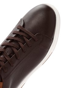 Primer plano de una zapatilla de cuero marrón con cordones a juego y suela de goma blanca sobre un fondo blanco.