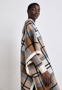 Poncho din amestec de lână cu un model mare în carouri în nuanțe de bej, maroniu și bleumarin, având margini ribbate și un design deschis în față.
