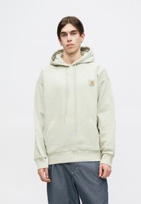 Sudadera color beis claro con un bolsillo delantero, capucha con cordón y puños acanalados. Presenta un pequeño parche de logo en la parte superior izquierda. Textura suave.