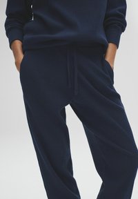 Persona vestida con pantalones de punto acanalado azul oscuro y un suéter a juego, con las manos en los bolsillos, frente a un fondo liso.