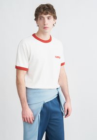 Λευκό βαμβακερό t-shirt με κόκκινη επένδυση, με μικρό κόκκινο λογότυπο. Ένα μπλε ύφασμα δεμένο γύρω από τη μέση ολοκληρώνει την εμφάνιση. Παντελόνια σε σκούρο μπλε χρώμα.