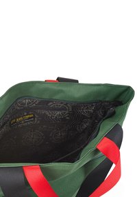 Bolsa de lona verde con correas rojas y negras. El interior presenta un forro negro con un patrón de brújula y logo. Diseño de apertura superior.