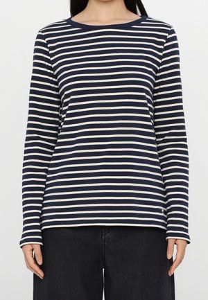 Kobieta ubrana w granatowo-białą bluzkę w paski z długim rękawem, w czarnych spodniach, stojąca na jednolitym białym tle.